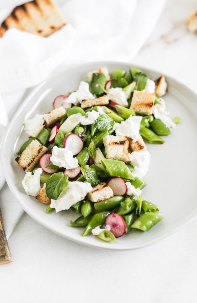 snap pea salad with burrata, radishes, mint and sourdough croutons on a white plate.
