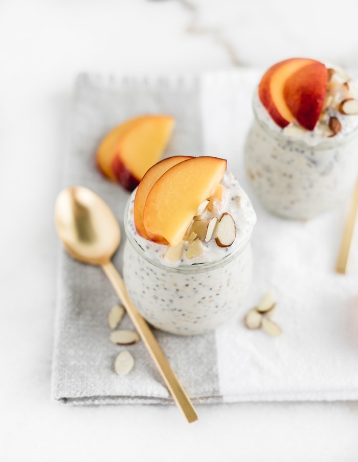 almond peach overnight oats in glass jars topped with peach slices with gold spoons beside them.