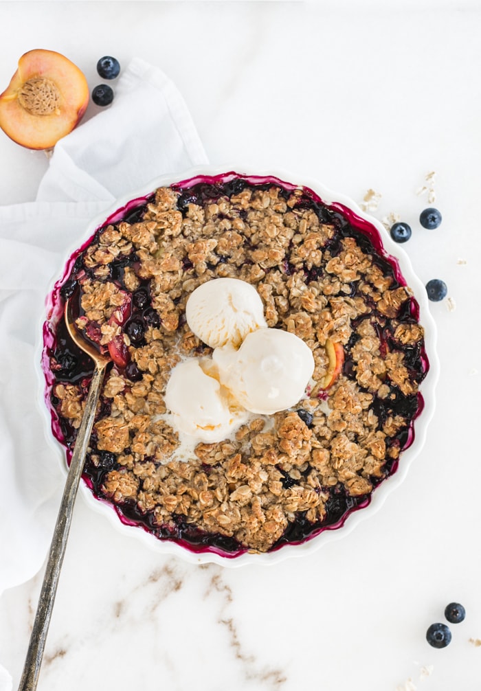 overhead view of blueberry peach crumble topped with ice cream in a round baking dish with a spoon in it.