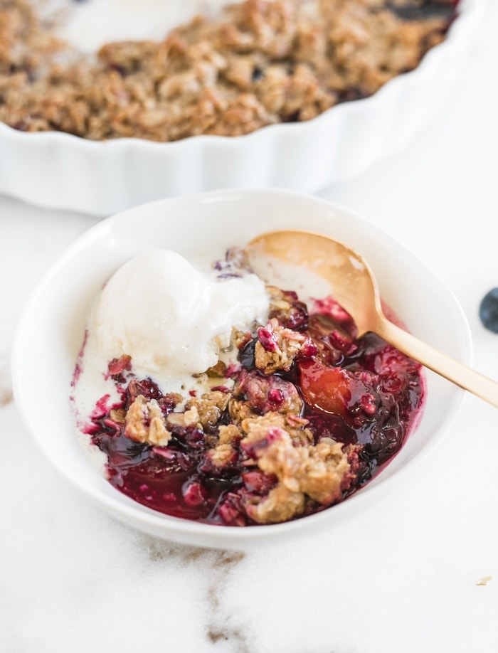 blueberry peach crumble in a bowl with ice cream and a gold spoon.