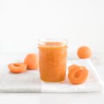 jar of low sugar apricot jam on a grey and white napkin surrounded by fresh apricot halves.