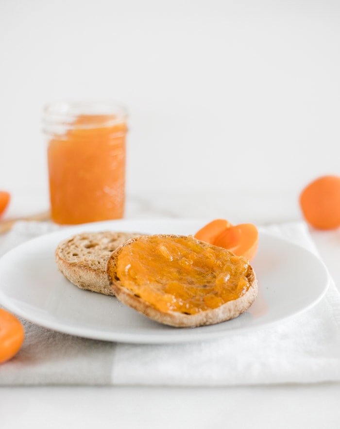 english muffin cut in half and spread with low sugar apricot jam on a white plate with a jar of jam in the background.