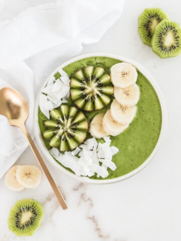 overhead view of pineapple kiwi green smoothie in a bowl topped with kiwi, banana slices and coconut flakes with a gold spoon beside it