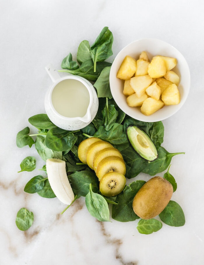overhead view of ingredients needed to make a pineapple kiwi green smoothie bowl on a white marble background.
