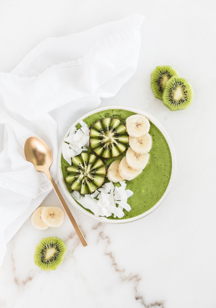 overhead view of pineapple kiwi green smoothie in a bowl topped with kiwi, banana slices and coconut flakes with a gold spoon beside it