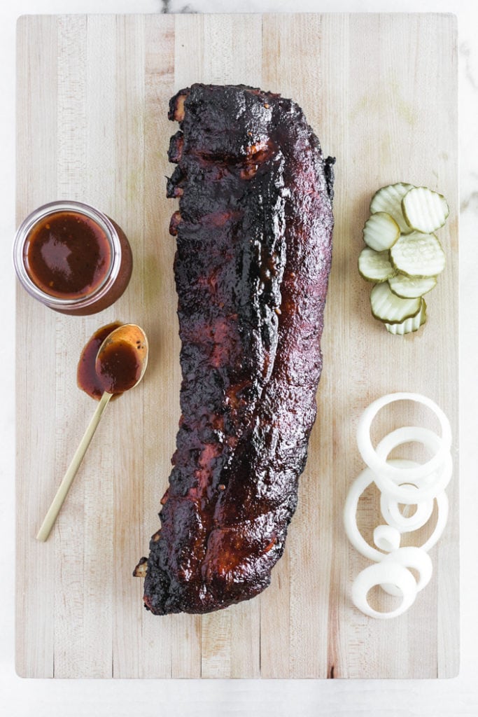overhead view of a rack of BBQ ribs cooked on the charcoal grill on a cutting board with pickles, onions and BBQ sauce