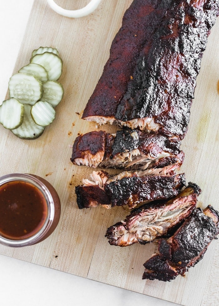 BBQ Ribs sliced on a cutting board with pickles and a jar of BBQ sauce.