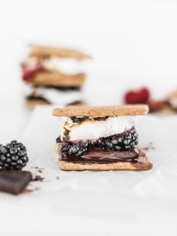 a dark chocolate berry s'more with melted chocolate and blackberries with stacked s'mores in the background