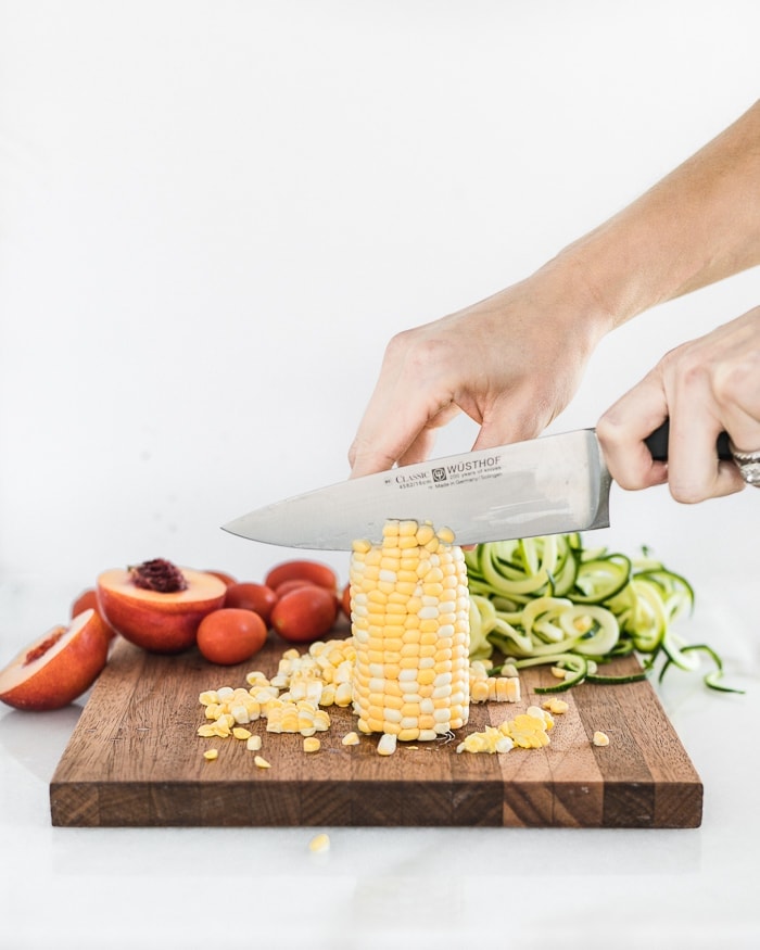 hands cutting corn off the cobb on a cutting board with zucchini noodles, cherry tomatoes, and peaches in the background