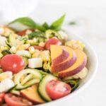 closeup of corn, zucchini noodles, tomato and mozzarella salad with sliced peach and basil on top in a white bowl