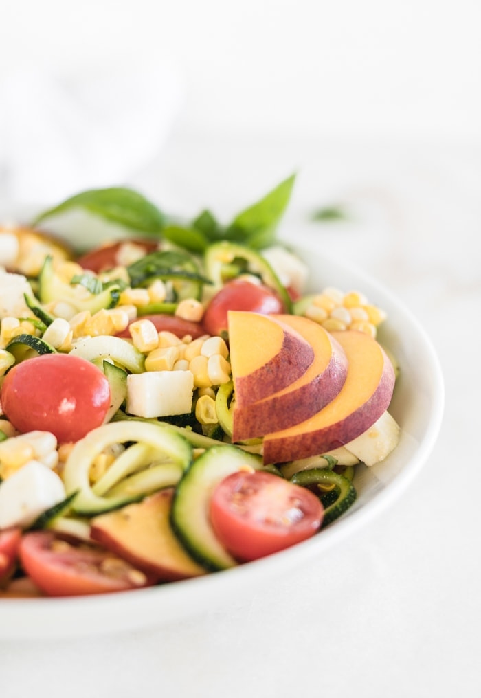 closeup of corn, zucchini noodles, tomato and mozzarella salad with sliced peach and basil on top in a white bowl