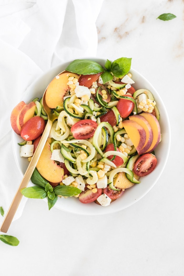 overhead shot of zucchini noodle, corn, peach and tomato caprese salad in a white bowl with a gold spoon