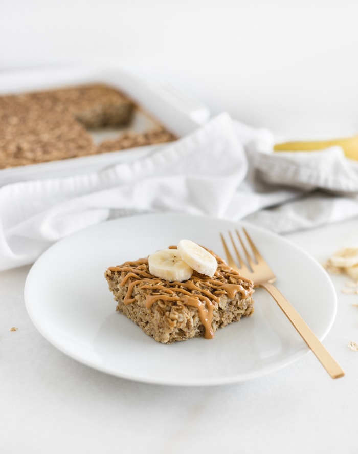 square of peanut butter banana baked oatmeal drizzled with peanut butter and topped with banana slices on a white plate with a gold fork.