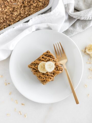 overhead shot of a square of peanut butter banana baked oatmeal drizzled with peanut butter and topped with banana slices on a white plate with a gold fork.