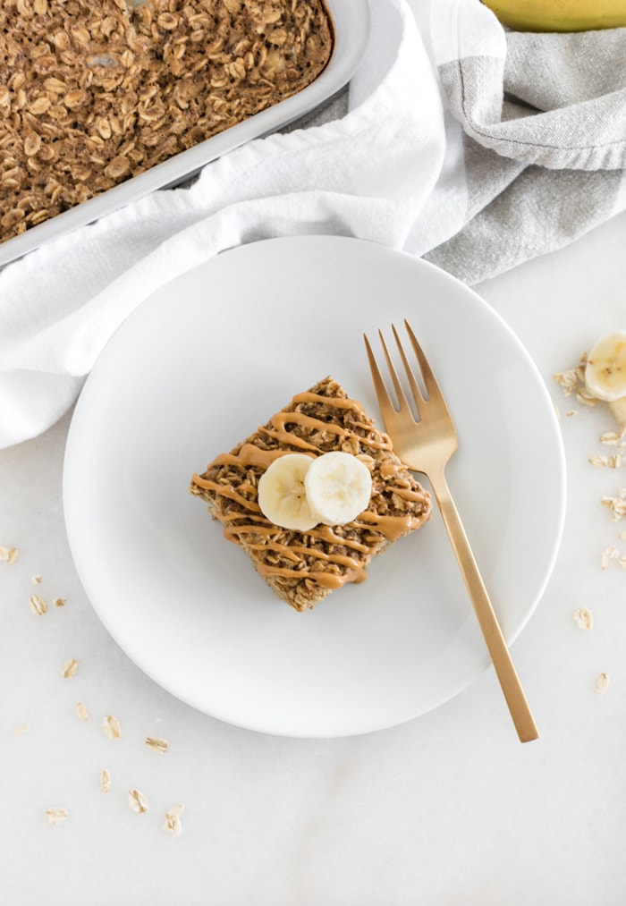overhead shot of a square of peanut butter banana baked oatmeal drizzled with peanut butter and topped with banana slices on a white plate with a gold fork.