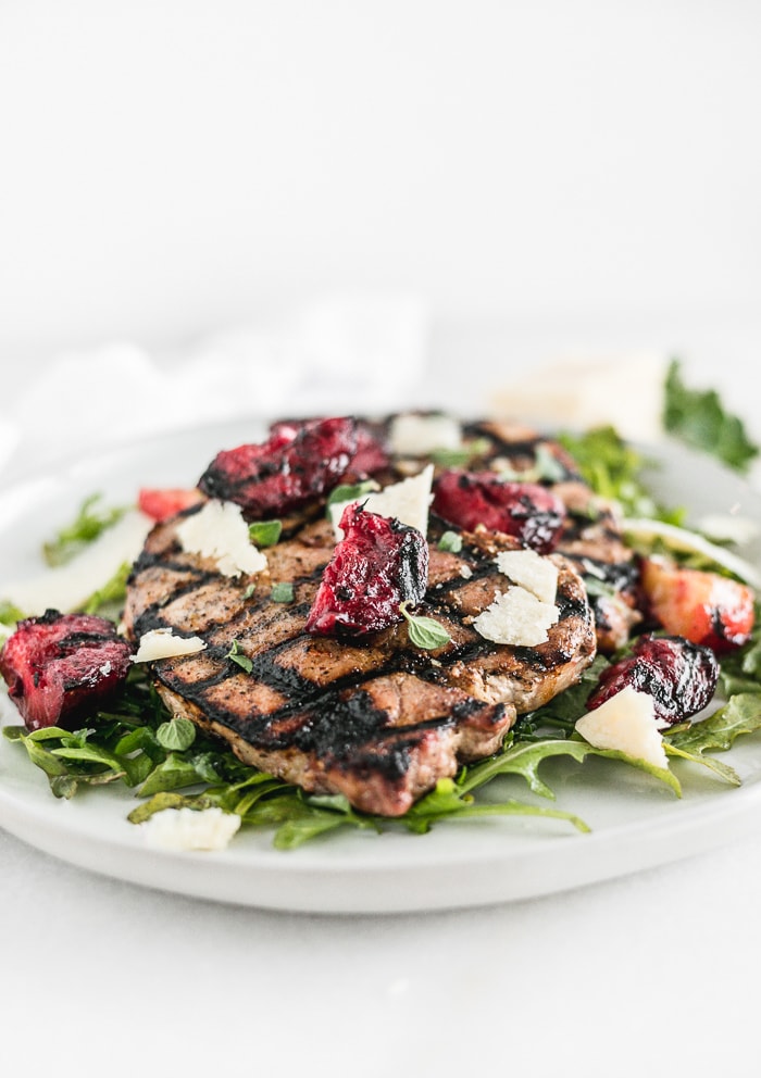 grilled pork chops on top of arugula with grilled plums and parmesan on top, on a white plate.