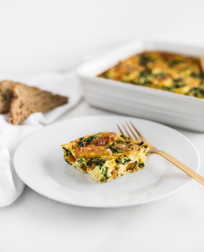 square of sweet potato and veggie breakfast casserole on a white plate with a gold fork and toast and a casserole dish in the background