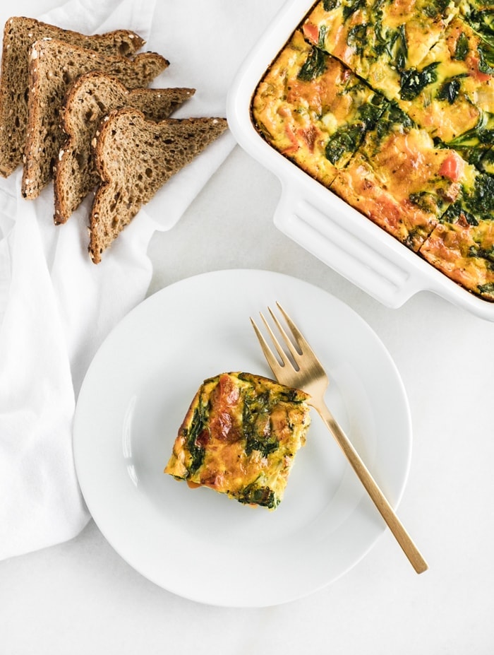 overhead shot of a square of veggie breakfast casserole on a white plate with a gold fork, with toast and the casserole dish