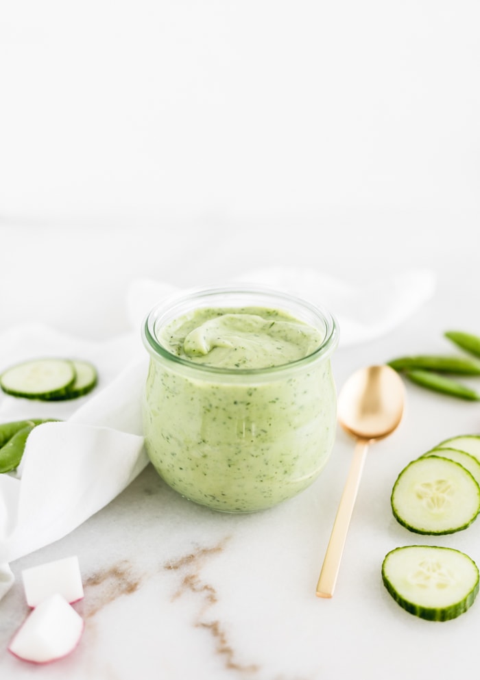 jar of avocado green goddess dressing with a gold spoon and sliced vegetables beside it