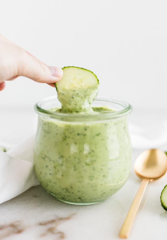 hand dipping a cucumber slice into avocado green goddess dressing in a jar.