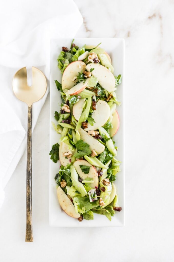 overhead shot of celery apple salad with walnuts and dates on a rectangle serving platter with a gold spoon.