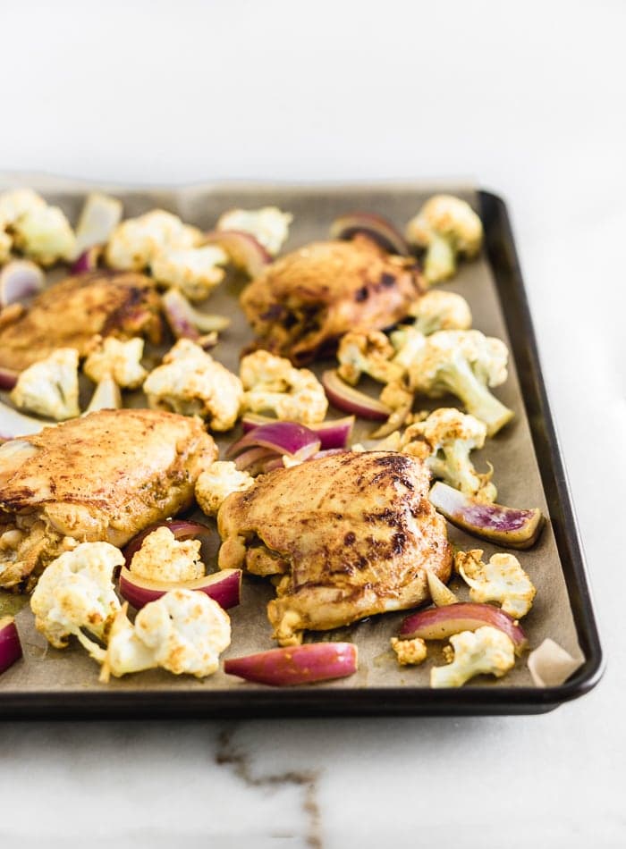 sheet pan with chicken thighs, cauliflower, and onions spread on it.