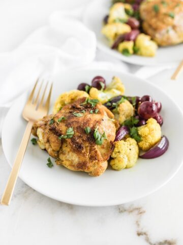 honey turmeric chicken thighs with roasted cauliflower and grapes on a white plate with a gold fork, with another plate of food in the background.