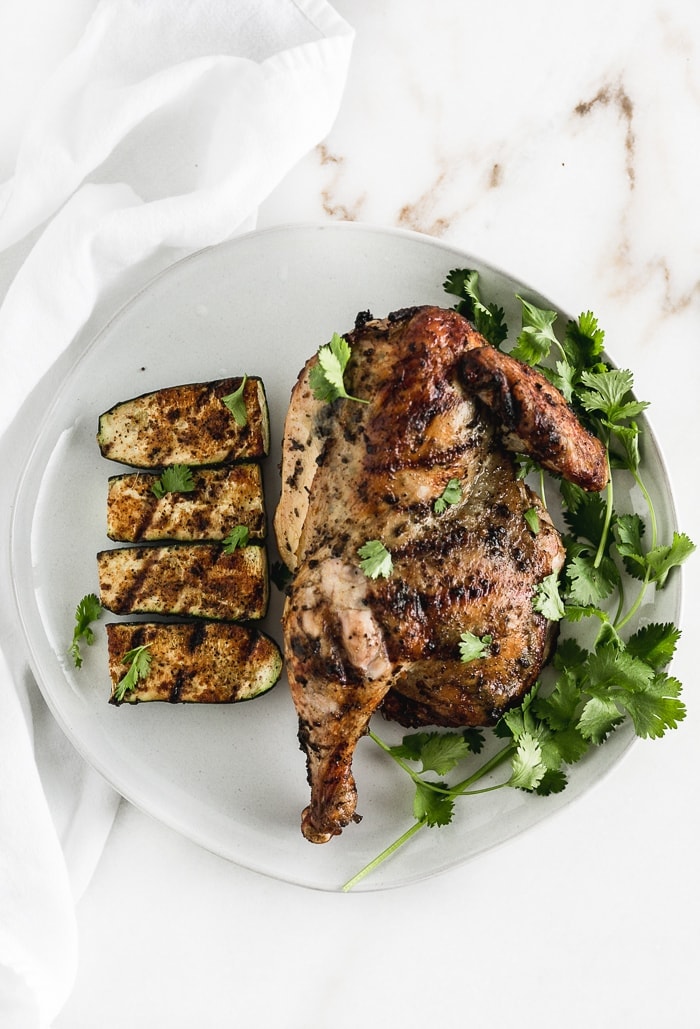 A half grilled chicken on a plate with cilantro and grilled zucchini.