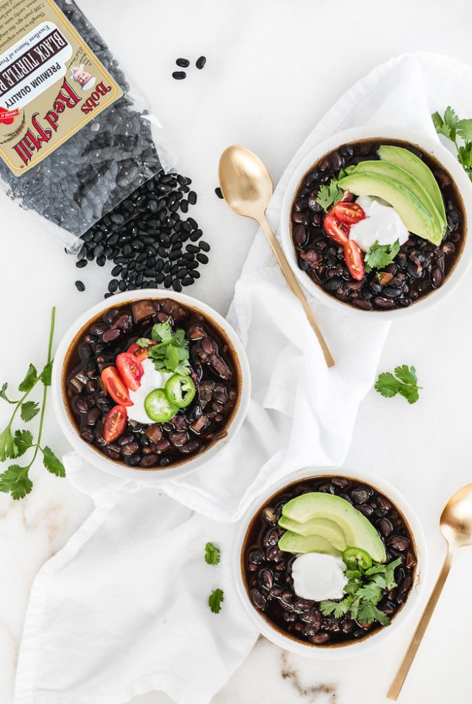 overhead view of three bowls of mexican black bean soup in white bowls surrounded by gold spoons, cilantro and a bag of black beans.