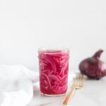 pickled red onions in a jar with a gold fork next to it and an onion in the background.