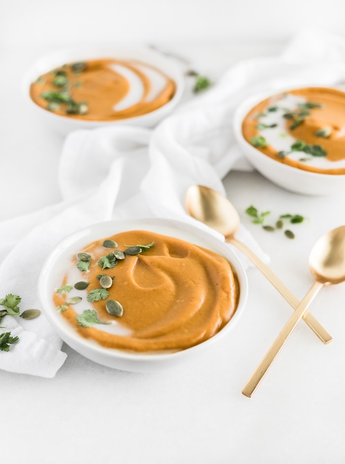 three bowls of coconut curry pumpkin soup topped with cilantro and pepitas with two gold spoons.