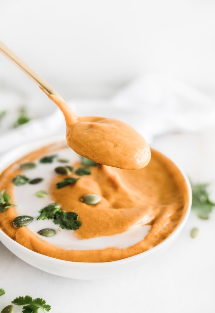 a gold spoon lifting a spoonful from a bowl of thai coconut curry pumpkin soup topped with coconut milk, cilantro and pepitas.