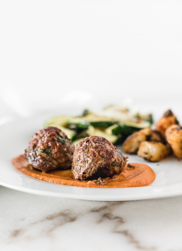 two meatballs on top of romesco sauce on a white plate with zucchini and potatoes in the background of the plate.