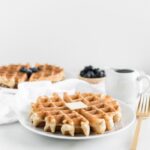 whole wheat waffle with a pat of butter on a white plate and a gold fork beside it with syrup, blueberries and another waffle in the background.