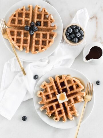 overhead view of thwo whole wheat buttermilk waffles on white plates with gold forks, a dish of syrup, and a bowl of blueberries.