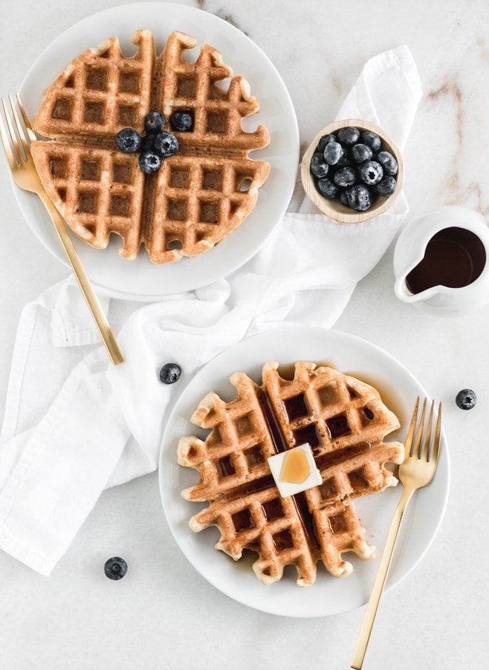 overhead view of thwo whole wheat buttermilk waffles on white plates with gold forks, a dish of syrup, and a bowl of blueberries.