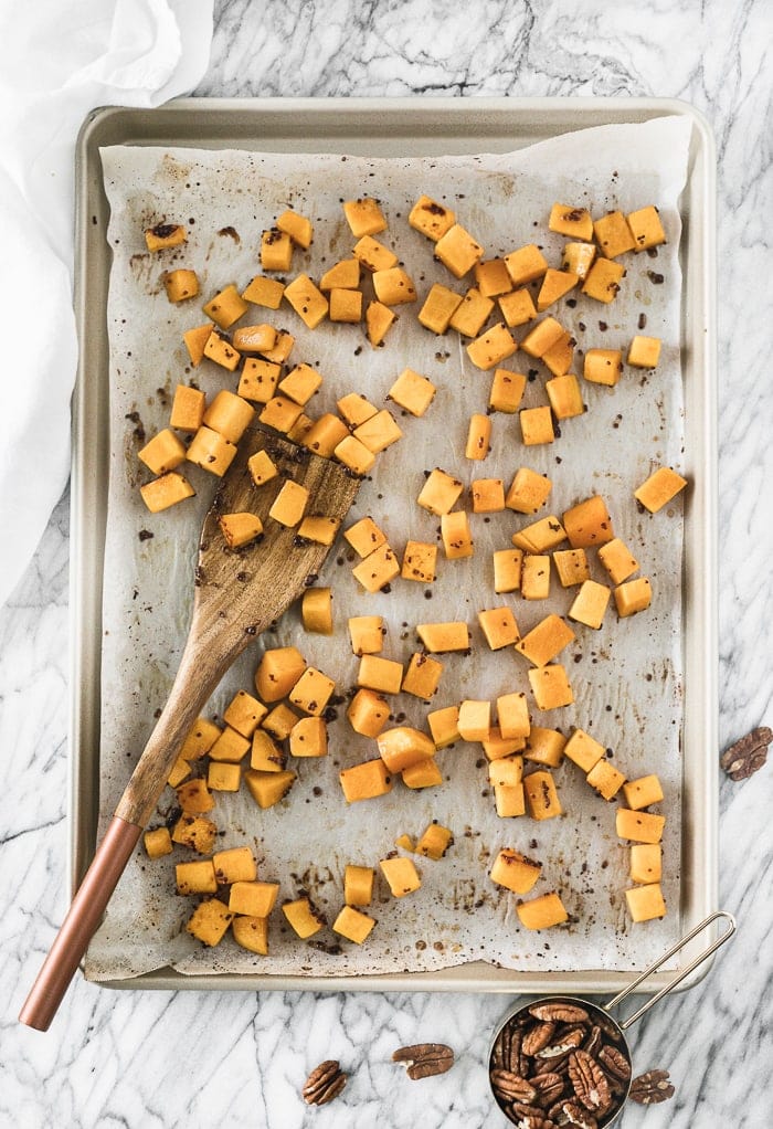 baking sheet with roasted butternut squash cubes and a wooden spatula on it surrounded by pecans.