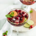 Glass of Christmas punch with cranberries, lime, orange slices and mint with another glass of punch in the background.