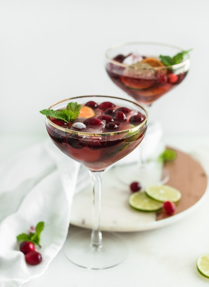 Two coupe glasses filled with red Christmas punch surrounded by sliced limes and cranberries.