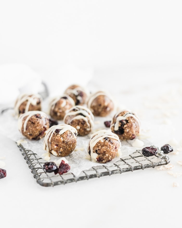 cranberry bliss balls drizzled with white chocolate on a rectangular cooling rack covered in parchment, surrounded by dried cranberries and white chocolate chips.