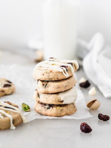 cranberry pistachio shortbread cookies with white chocolate stacked on top of each other with cranberries and pistachios around them.