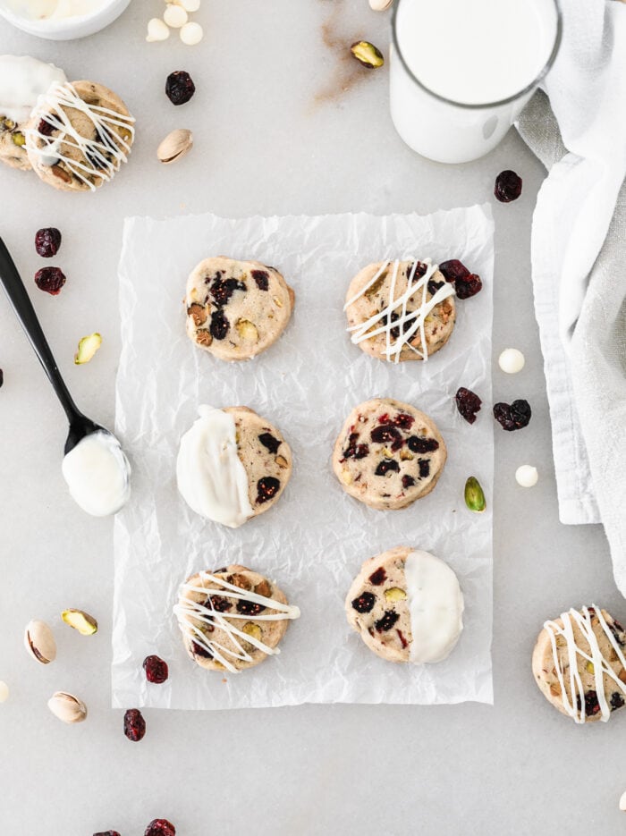 overhead view of cranberry pistachio shortbread cookies on a piece of parchment surrounded by a spoon dipped in melted white chocolate, cranberries, pistachios, white chocolate chips and a glass of milk.