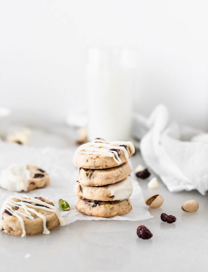 four stacked cranberry pistachio cookies with white chocolate surrounded by cranberries, pistachios, and more cookies.