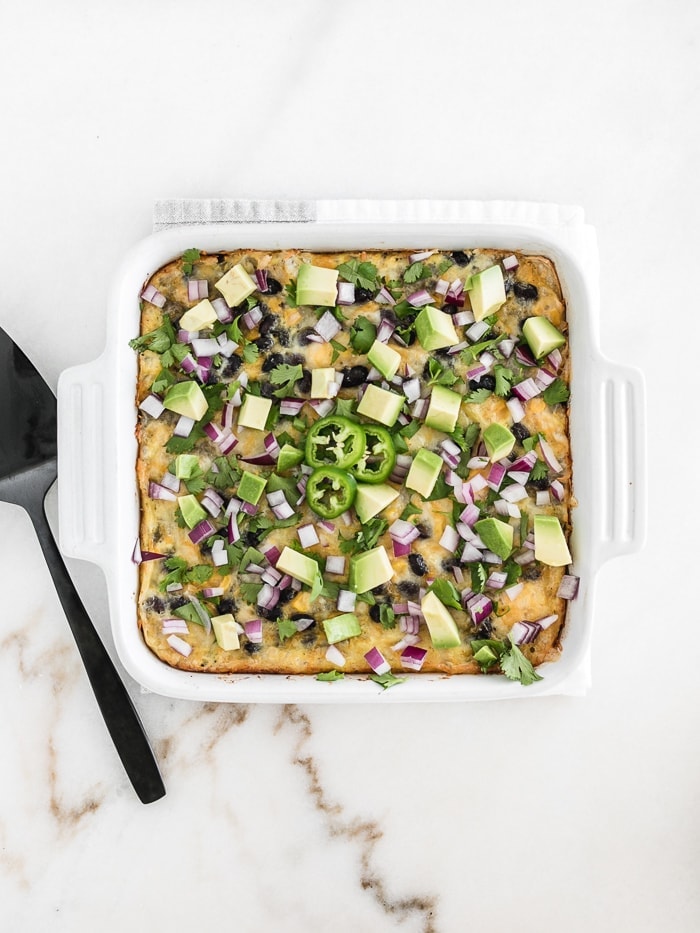 Overhead view of mexican breakfast casserole topped with avocado, cilantro and red onion in a white baking dish with a black pie server beside it.