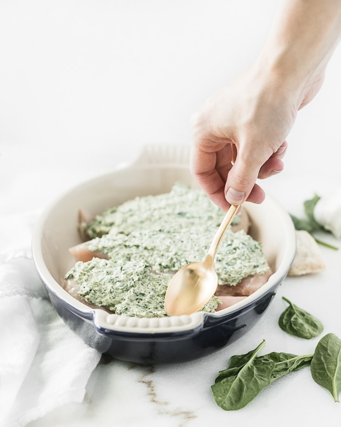 hand using a gold spoon to spread spinach artichoke topping on top of chicken breasts in a blue oval baking dish.