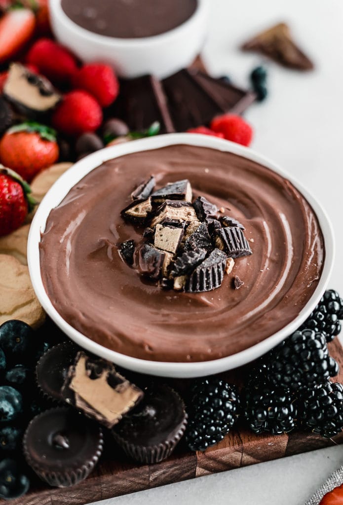closeup of chocolate peanut butter dip in a white bowl topped with chopped peanut butter cups and surrounded by berries and chocolate.