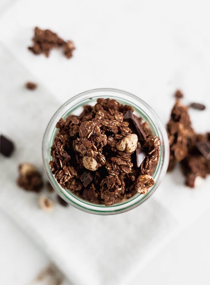 overhead view of chunky chocolate hazelnut granola in a glass jar surrounded by granola pieces.