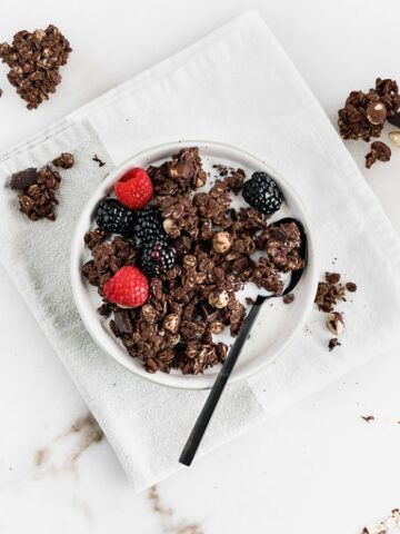 overhead shot of chocolate hazelnut granola in a bowl with milk and berries with a black spoon on top of a white and grey napkin.