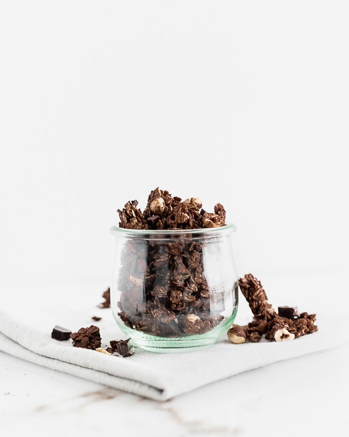 chunky chocolate hazelnut granola in a glass jar surrounded by granola pieces.