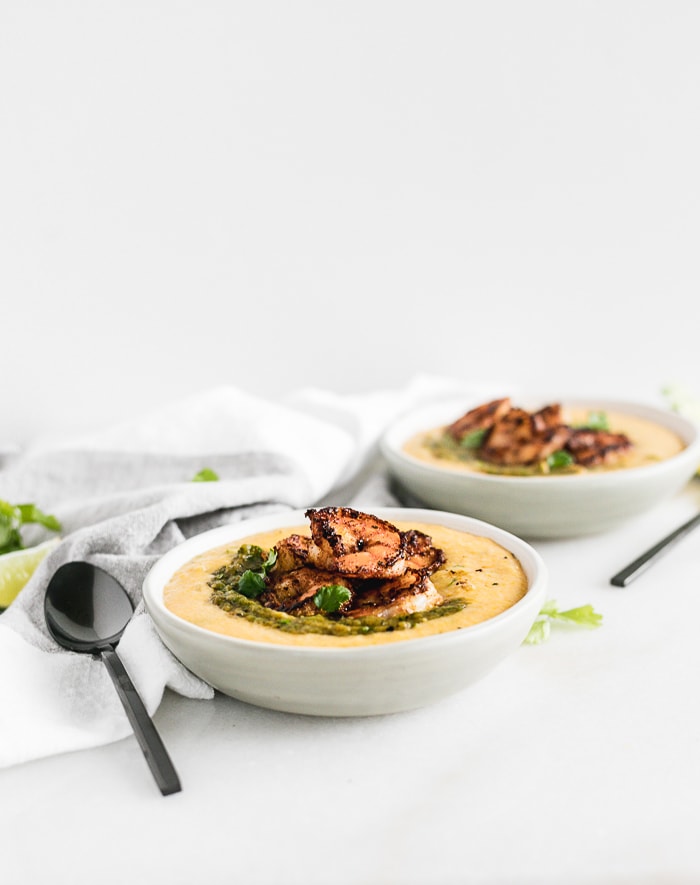 two bowls of green chile shrimp and grits with black spoons next to them.